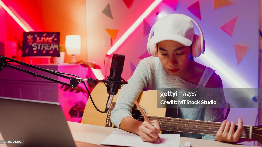 Happy Asian girl influencer wear headphone record podcast on laptop play guitar writing notes to music book onair online live streaming at studio room.