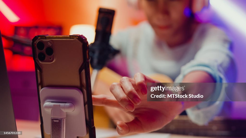 Close-up Happy Asian girl influencer wear headphone with guitar record podcast onair online live streaming talk follower comment and audience on phone at home studio.