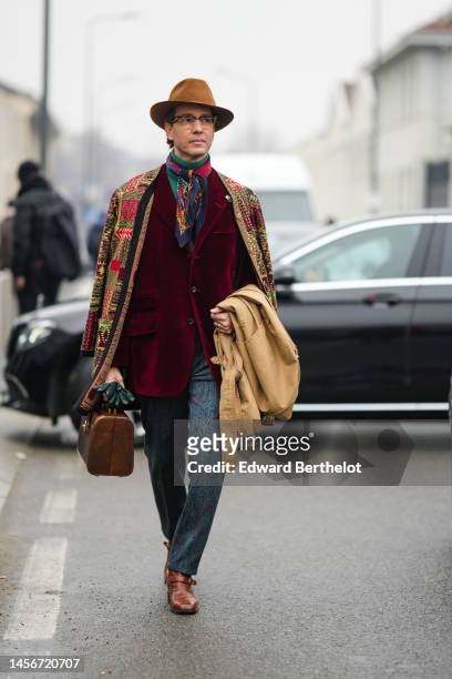 Guest wears a brown felt hat, glasses, a green ribbed wool turtleneck pullover, a multicolored pleated scarf, a burgundy velvet blazer jacket, a...