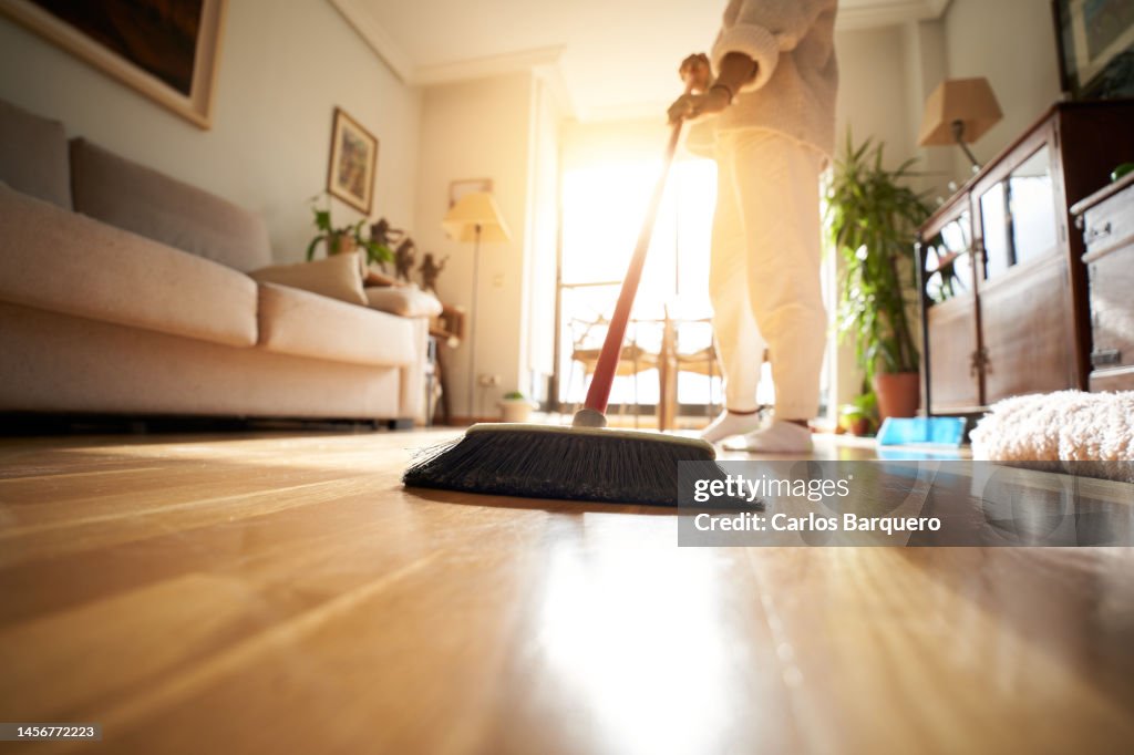 Unrecognizable woman sweeping the parquet floor, window light reflection.