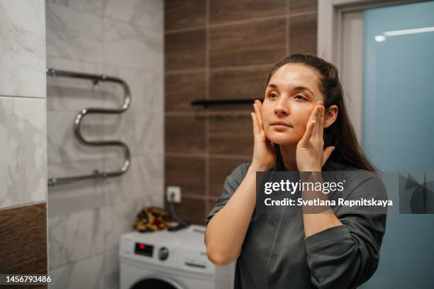 woman applying cream on her face in front of a mirror - masaje facial fotografías e imágenes de stock
