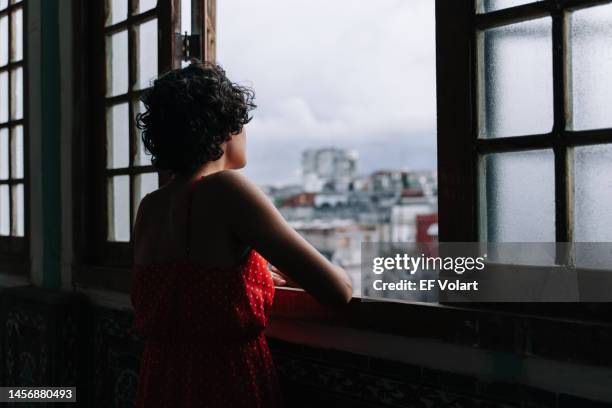 pensive young latin girl contemplating the city through window at home - open grave - fotografias e filmes do acervo