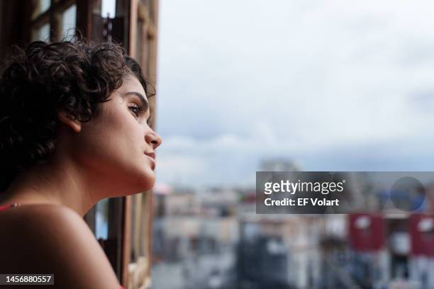 young pensive hispanic woman looking the city through window at home - greater antilles stock pictures, royalty-free photos & images