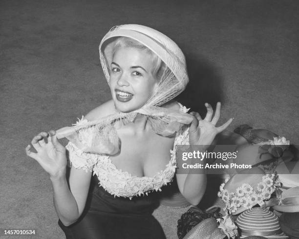 American actress, singer and glamour model Jayne Mansfield , both hands of the ribbons of her bonnet, wearing a low-cut dress beside a display of...