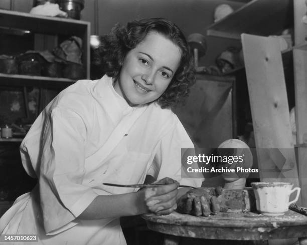 British-American actress Olivia de Havilland takes a break from her hobby of sculpting, a sculpted clay head beside her in her workshop, United...