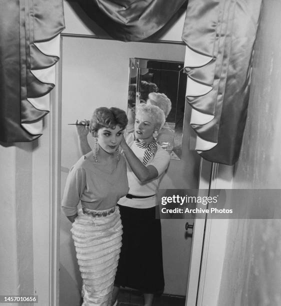 American actress Debra Paget, wearing long drop earrings, her hands behind her back, as a hairdresser adjusts her hair, July 1954.