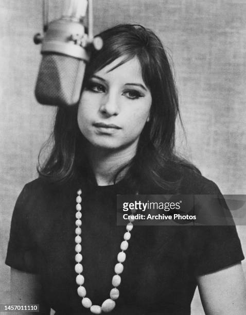 American singer and actress Barbra Streisand wearing a black short sleeve top with a white beaded necklace, standing before microphone in an...