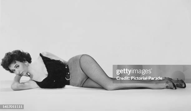 Puerto Rican actress, singer and dancer Rita Moreno, wearing a black scoop neck top with hotpants and fishnet stockings, leaning on her right elbow...