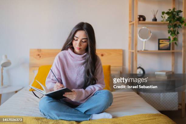 frontal view of a young woman with long dark hair, sitting on the bed in her room writing in a notebook. - non moving activity stock pictures, royalty-free photos & images