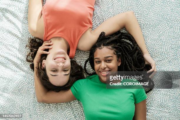 couple of young women stretched out in bed touching their hair looking at camera. couple and women concept - depth of field togetherness looking at the camera ストックフォトと画像