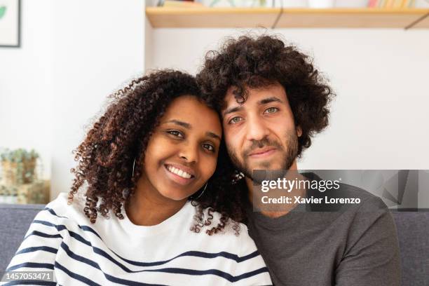 multiracial portrait of young adult couple looking at camera sitting on sofa at home - interracial wife photos stock-fotos und bilder