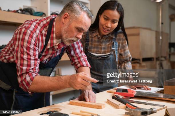 craftsman senior man teaching woman apprentice at woodshop, carpenter training wood for female standing at wood table in workshop, national carpenters day - nationaltrainer stock pictures, royalty-free photos & images