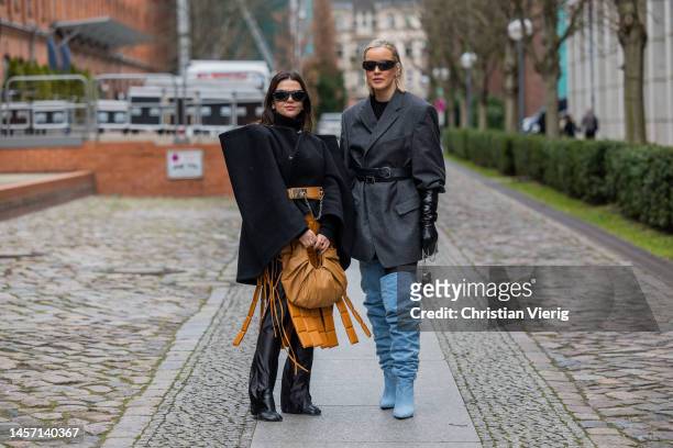 Amelie Stanescu wears brown ripped skirt, belt, black cape, brown bag, black pants, boots, sunglasses & Tina Haase wears grey belted blazer, blue...