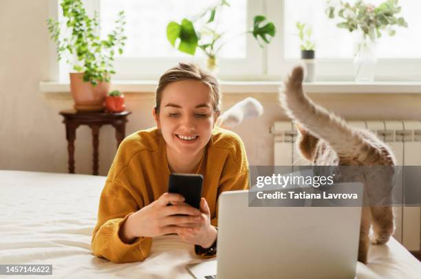 young attractive female in yellow cardigan studying with her cat. she is using smartphone and laptop. distance education, leisure activities at home, surfing net concept - cat laptop stockfoto's en -beelden