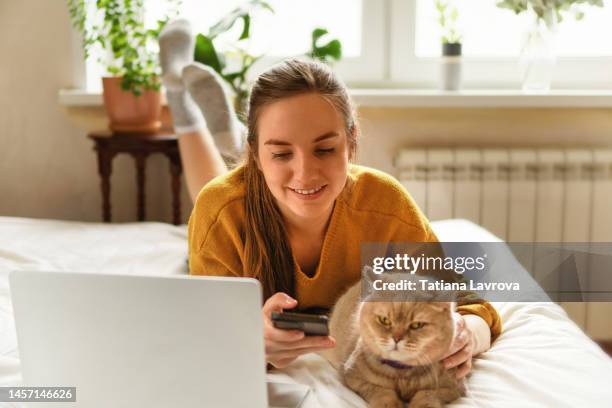 young attractive female in yellow cardigan studying with her cat. she is using smartphone and laptop. distance education, leisure activities at home, surfing net concept - portrait chantier stock-fotos und bilder