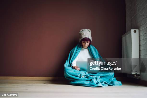 woman wrapped in a blanket working and online shopping on a sick leave near cold radiator at home. copy space - frost stock photos et images de collection