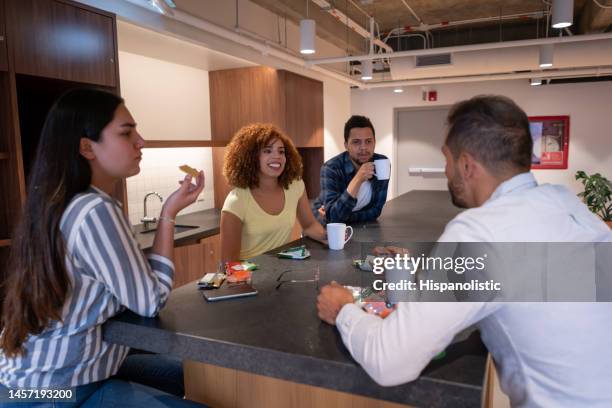 young team at a coworking office taking a break eating snacks and enjoying coffee while talking and smiling - office snacks stock pictures, royalty-free photos & images