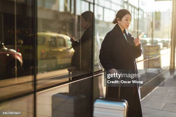 young asian business woman with suitcase, ordering taxi with smartphone on city street - walk new york stock pictures, royalty-free photos & images