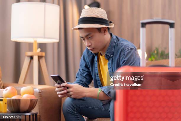 ready to travel abroad,asia adult male man casual cloth sit on sofa couch next to luggage travel bag, hand reserve booking hotel room and flight ticket by online application smart device east travel - yuri malenchenko stock pictures, royalty-free photos & images