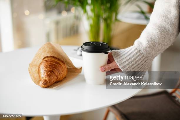 female hand holding a disposable glass with coffee and fresh croissant. concept of delivery food - coffe to go stockfoto's en -beelden
