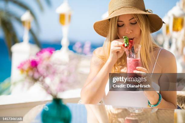 young blonde woman sitting in the beach restaurant drinking a red cocktail with the turquoise sea  in the background - blue white summer hat background stock pictures, royalty-free photos & images