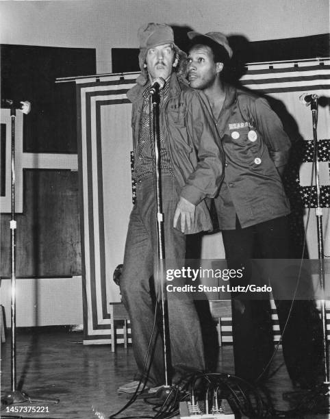 Full-length shot of Donald Sutherland and Paul Mooney, wearing US Army shirts and standing on stage behind a microphone, participating in an...