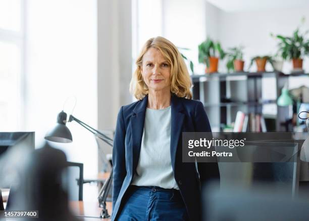 portrait of a confident mature businesswoman standing by a desk in office - portrait work stockfoto's en -beelden