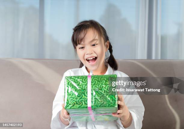 little asian girl smile and excited and holding red gift box on sofa in living room background.child holding gift box in christmas and new year.asian child girl toddler baby smile and surprise. - the red room opening party inside stock pictures, royalty-free photos & images