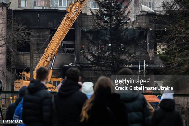 Locals look over the fence at the removing of a preschool building rubble after a helicopter crashed on its territory on January 18, 2023 in Brovary,...