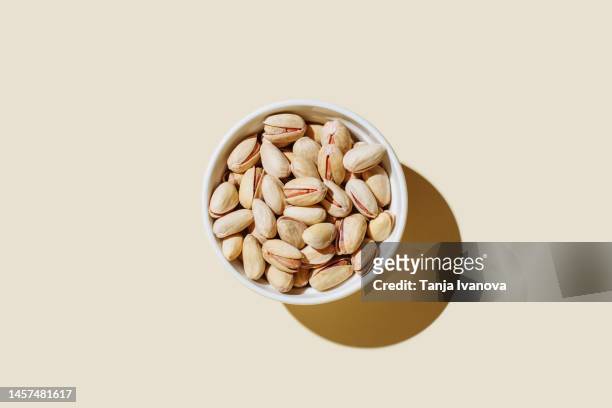 pistachio nuts in white bowl isolated on beige background. minimal flat lay style. top view, copy space. - pistachio stock pictures, royalty-free photos & images