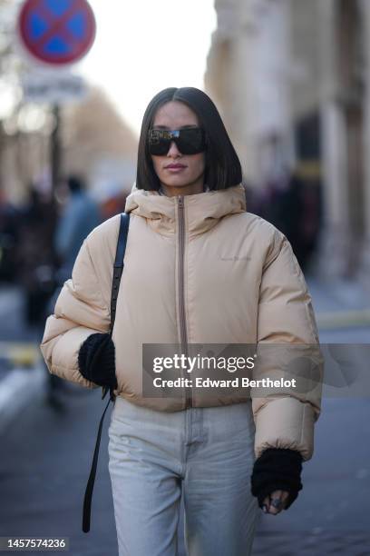 Alexandra Guerain wears black large futurist sunglasses, a beige zipper high neck / puffer jacket from Acne Studio, a black shiny leather shoulder...