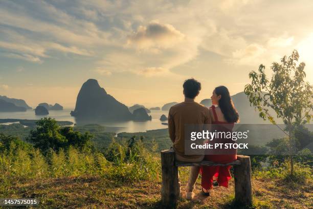 man and woman in red dress sit on wooden chairs in view of ao samet nang chee phang nga with sea and mountains in phang nga province, thailand. - red sea rain stock pictures, royalty-free photos & images