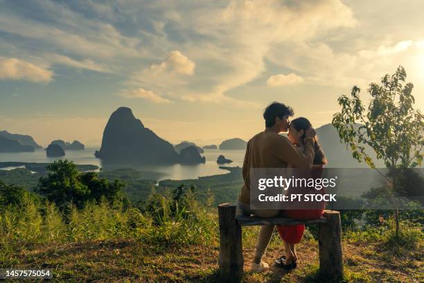 man and woman in red dress sitting on wooden chair and man kissing woman's forehead in view of ao samet nang chee phang nga bay with sea and mountains in phang nga province, thailand - red sea rain stock pictures, royalty-free photos & images