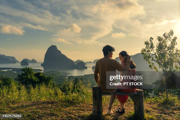 man and woman in red dress sitting on wooden chair and man stroking woman's hair in view of ao samet nang chee phang nga bay with sea and mountains in phang nga province, thailand - red sea rain stock pictures, royalty-free photos & images