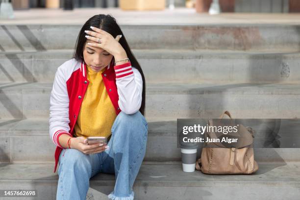 desperate teen student sitting on stairs while receiving school bullying via cellphone - cyberbullying stockfoto's en -beelden