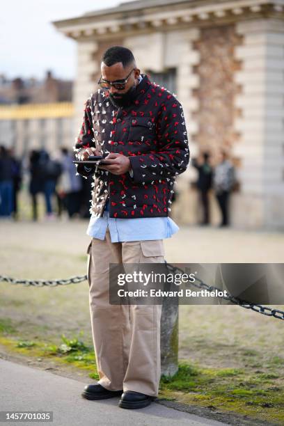 Guest wears black sunglasses, a pale blue striped print pattern shirt, a black with embroidered red and white pattern puffer jacket, beige cargo...