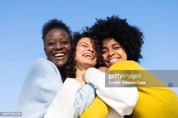 three cheerful young multi-ethnic women embracing on a blue background - female friendship stock pictures, royalty-free photos & images
