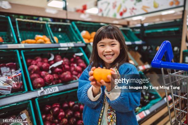 lovely little girl smiling at the camera joyfully while holding a little pumpkin in supermarket - supermarket uk stock pictures, royalty-free photos & images