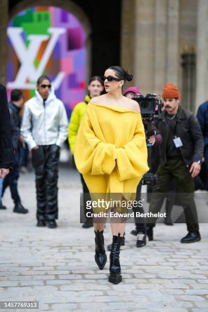 Rosalia wears sunglasses, a yellow wool oversized worn as an off-shoulder mini dress from Raf Simons, black high heeled pointed shiny boots, outside...