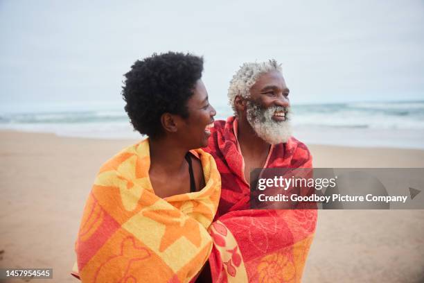 mature couple laughing while walking on a beach wrapped in towels after swimming - beach towel stock pictures, royalty-free photos & images