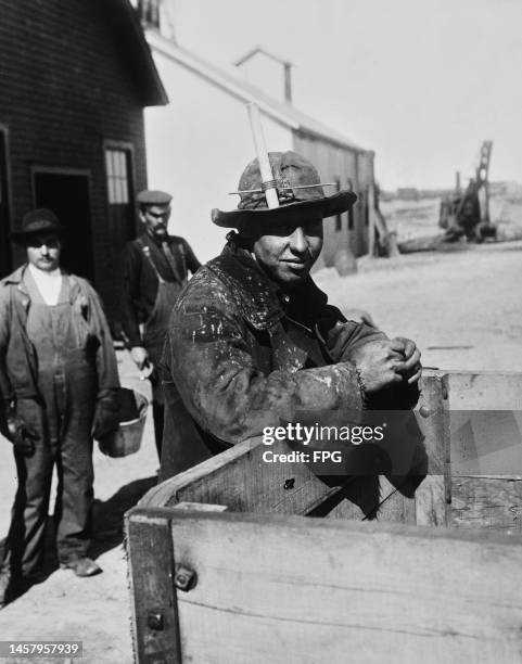 An iron ore miner leaning on the back of a cart, with two miners in the background, at the Mesabi Iron Range, Minnesota, circa 1905. Collectively...
