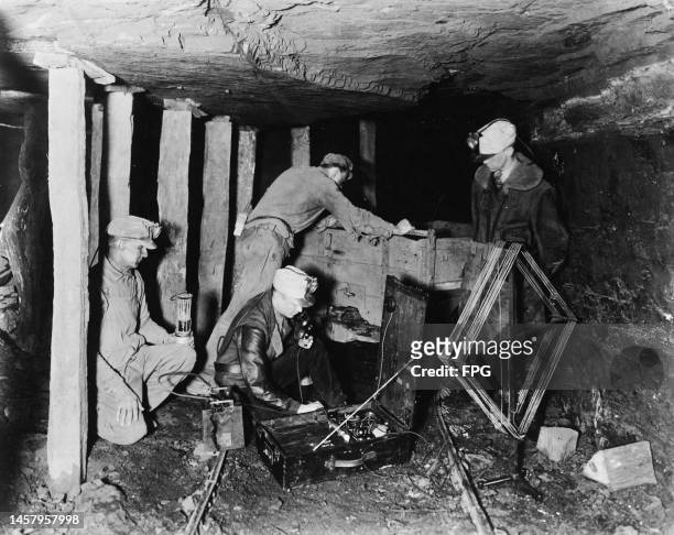 Coal miners wearing caps fitted with miners' lamps, and one sits on the mine floor to use a field telephone bedside a mining cart, one man sits...