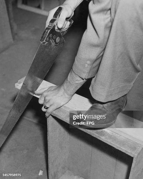 Partial view of a man using his foot to secure a length of wood, supported on a crate, as he saws on an construction site, United States, circa 1935.