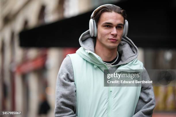 Guest wears gray headphones, a pale green zipper / sleeveless puffer jacket from Rains, a pale gray hoodie sweater, outside Rains, during Paris...