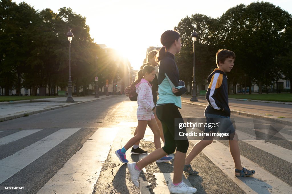 Group of people made up of children and adults crossing the street on the crosswalk, in the background there are trees and public lighting.