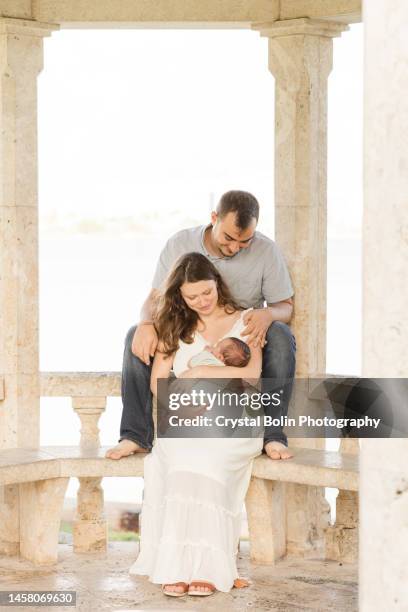 a sweet young family of three snuggled together in a gazebo on the water in a relaxing meditation garden in palm beach, florida for lifestyle newborn family photos - rock baby sleep stock pictures, royalty-free photos & images