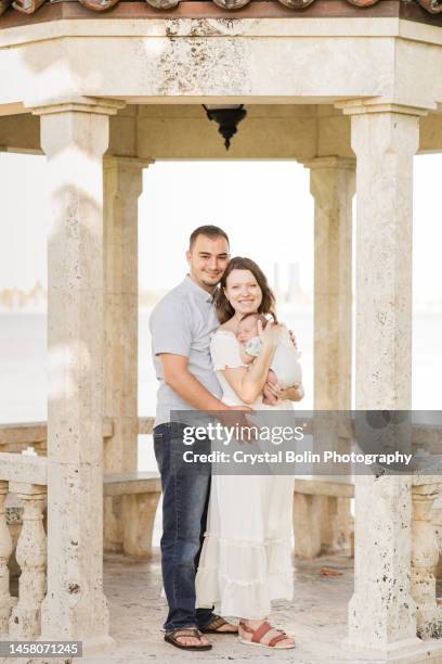 a sweet young family of three snuggled together in a gazebo on the water in a relaxing meditation garden in palm beach, florida for lifestyle newborn family photos - rock baby sleep stock pictures, royalty-free photos & images