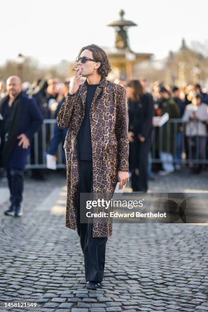 Guest wears black sunglasses, a black t-shirt, a brown and beige leopard print pattern long coat, black suit pants, black shiny leather shoes ,...