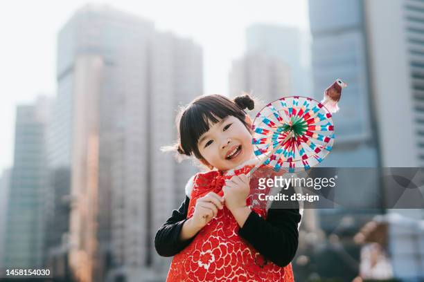 portrait of a lovely little asian girl in chinese costume holding a colourful windmill, smiling at camera joyfully, standing against urban city skyline in the city. celebrating chinese new year. traditional chinese culture. festival and celebration theme - traditional festival bildbanksfoton och bilder