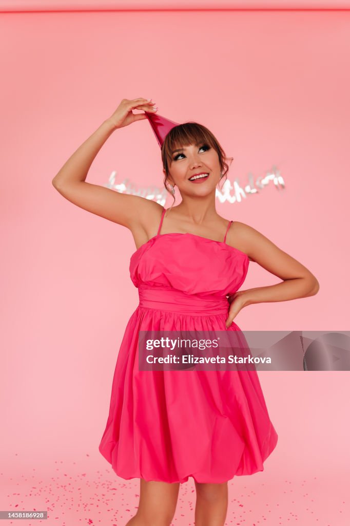A smiling happy woman holds balloons in the form of numbers and celebrates her thirtieth birthday in a bright pink dress on a colored pink background decorated with confetti. A cheerful girl in fashionable clothes and a festive hat has fun and celebrates
